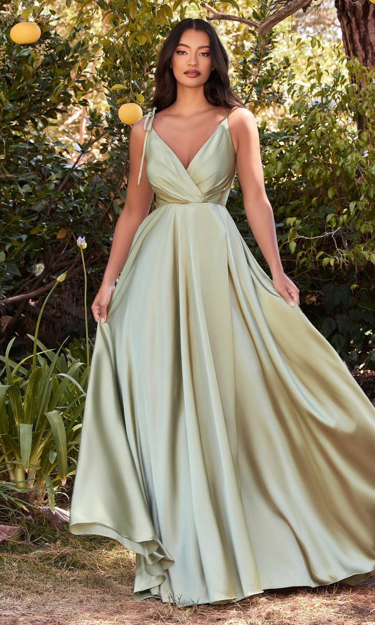 A woman stands outdoors among trees and plants, holding the skirt of her Shoulder-Tie Long Satin V-Neck Prom Dress BD105, a light green gown with an elegant v-neckline.